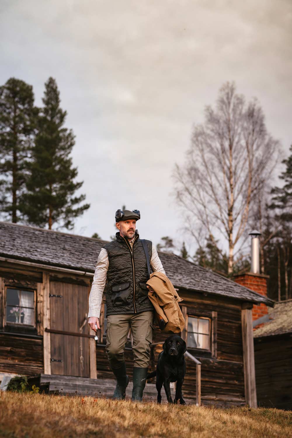 A man wearing a black vest, beige shirt, and green pants from Pinewood walks in front of a rustic wooden cabin with a black dog beside him