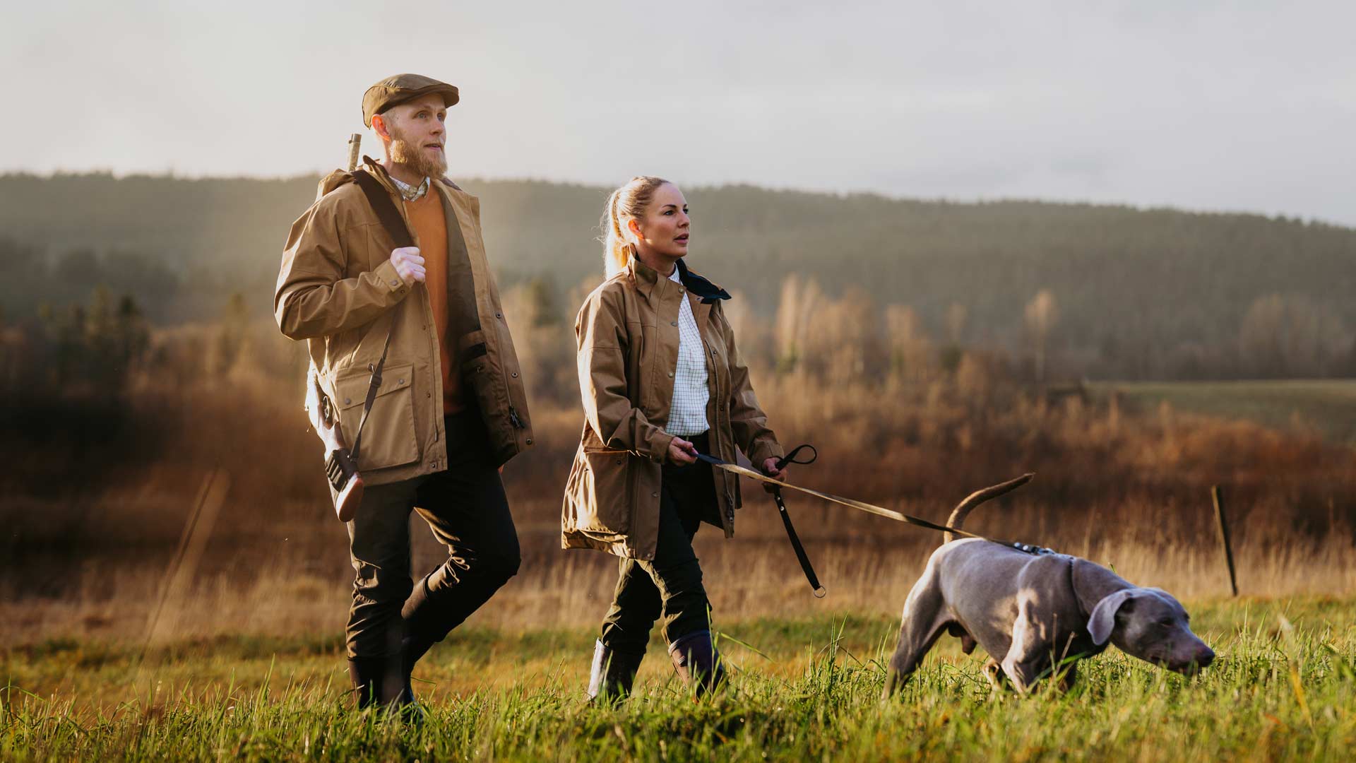 A man and a woman walking in a grassy field with a gray dog on a leash, wearing hunting gear from Pinewood
