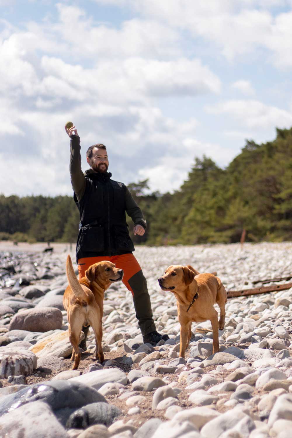 A man stands on a rocky beach holding up a treat for two dogs, wearing a black puffer vest over a dark shirt and bright orange shorts, with trees and a cloudy sky in the background.