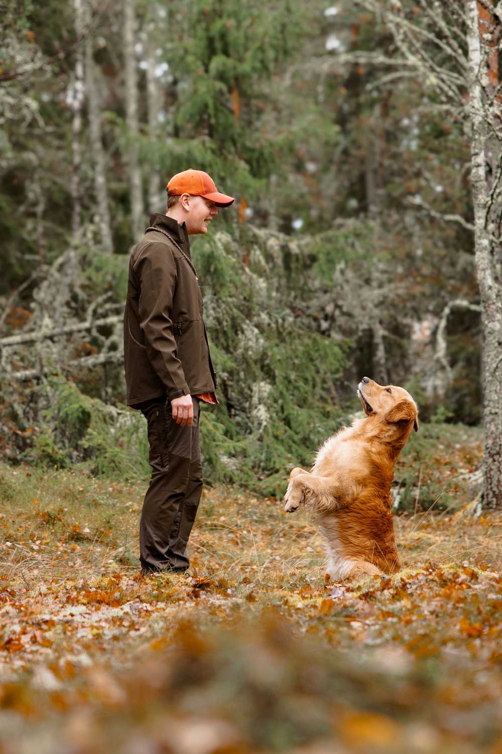 A man in a brown jacket and orange cap stands in a wooded outdoor setting next to a golden retriever dog sitting on a ground covered with fallen leaves.