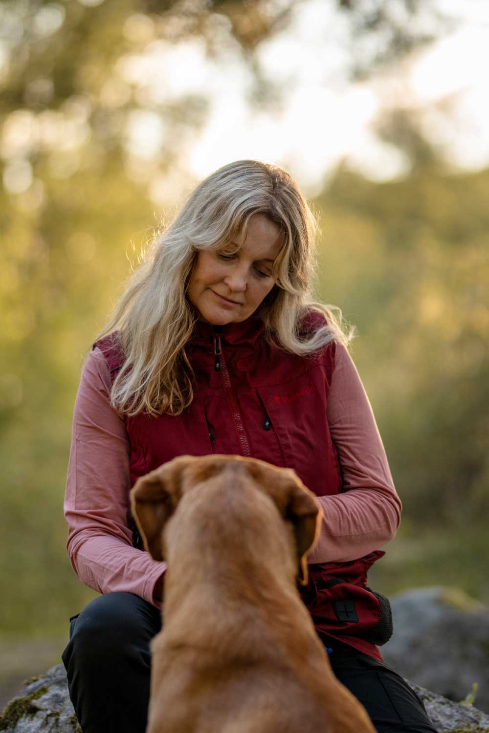A woman in a red puffer vest and pink long-sleeve shirt sits outdoors with a brown dog in the foreground, set against a soft-focus natural background.