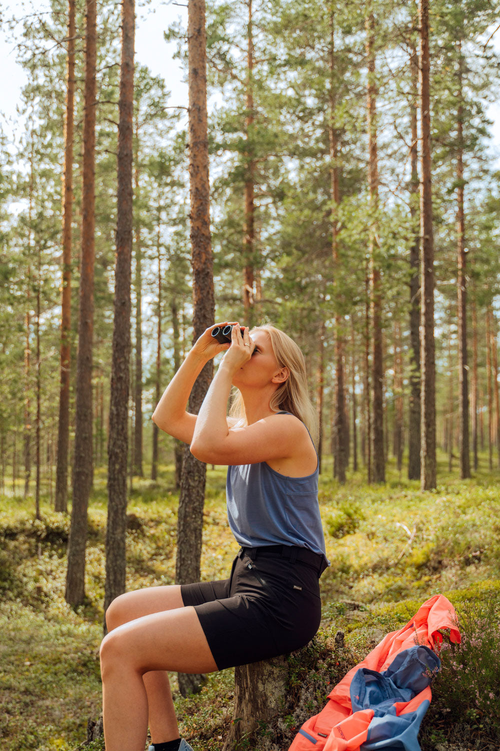 A woman is sitting on a tree stump in the forest, looking up at the sky with binoculars. She is wearing a tank top and hiking shorts.