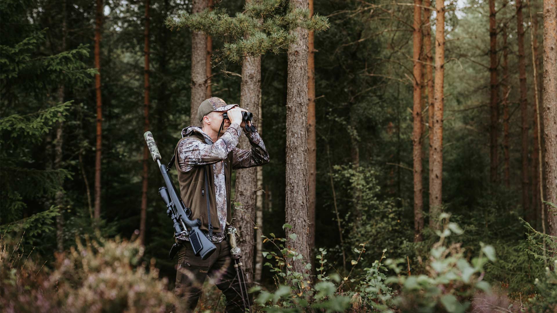 A man in camouflage hunting clothing from Pinewood stands in a dense pine forest, holding binoculars to his eyes with a rifle slung over his shoulder.