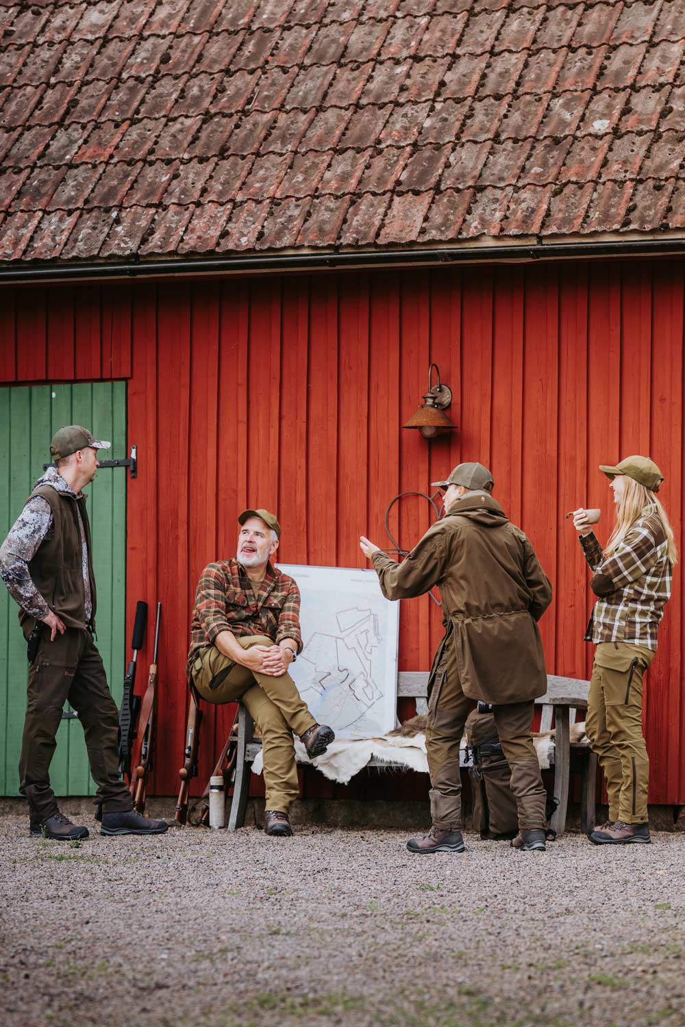 Four people dressed in hunting gear from Pinewood stand and sit in front of a red wooden barn with a tiled roof. One person holds a large white sketch or poster board, while others converse nearby in a staged outdoor scene.