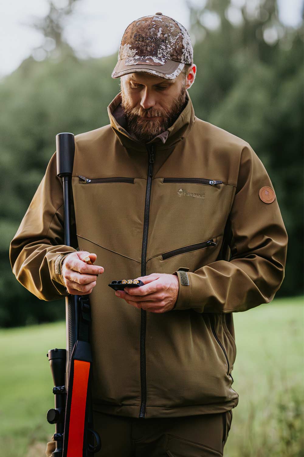 A bearded man wearing a brown camouflage cap and a brown hunting jacket from Pinewood stands outdoors holding a shotgun with a blurred green field and trees in the background.