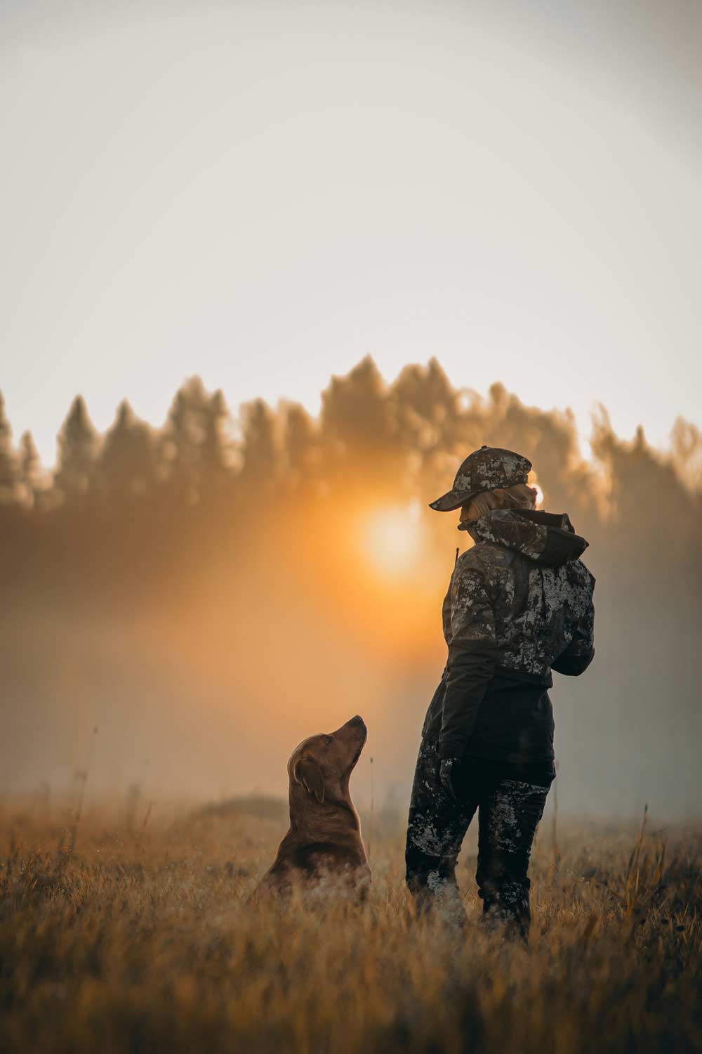 A person in camouflage hunting clothing  from Pinewood stands in a grassy field at sunset with a dog beside them, silhouetted against a warm glowing sky and tree line.