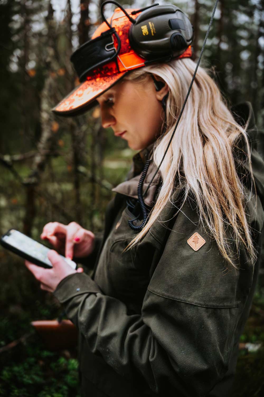 A woman in a forest wearing an orange camouflage hunting hat black over-ear headphones, and an olive green hunting jacket from Pinewood looking down at a phone.